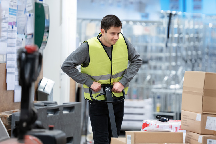 A man wearing a yellow vest pushes some boxes on a trolley in a warehouse.
