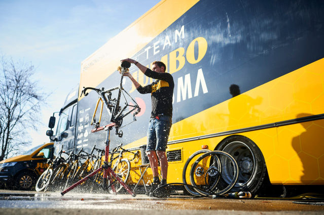 A mechanic works on a bicycle in the sunlight in front of the Team Jumbo-Visma bus.