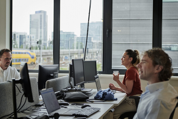 Colleagues sit talking at their desks next to a large window overlooking a city.