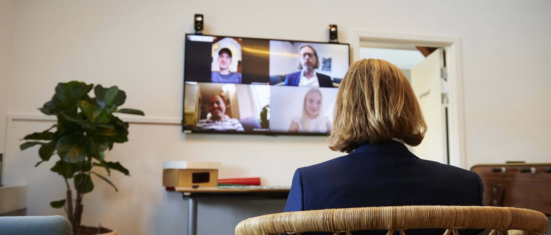 A man with long blonde hair wearing a suit sits in a chair facing some attendees in a video call on the big screen.