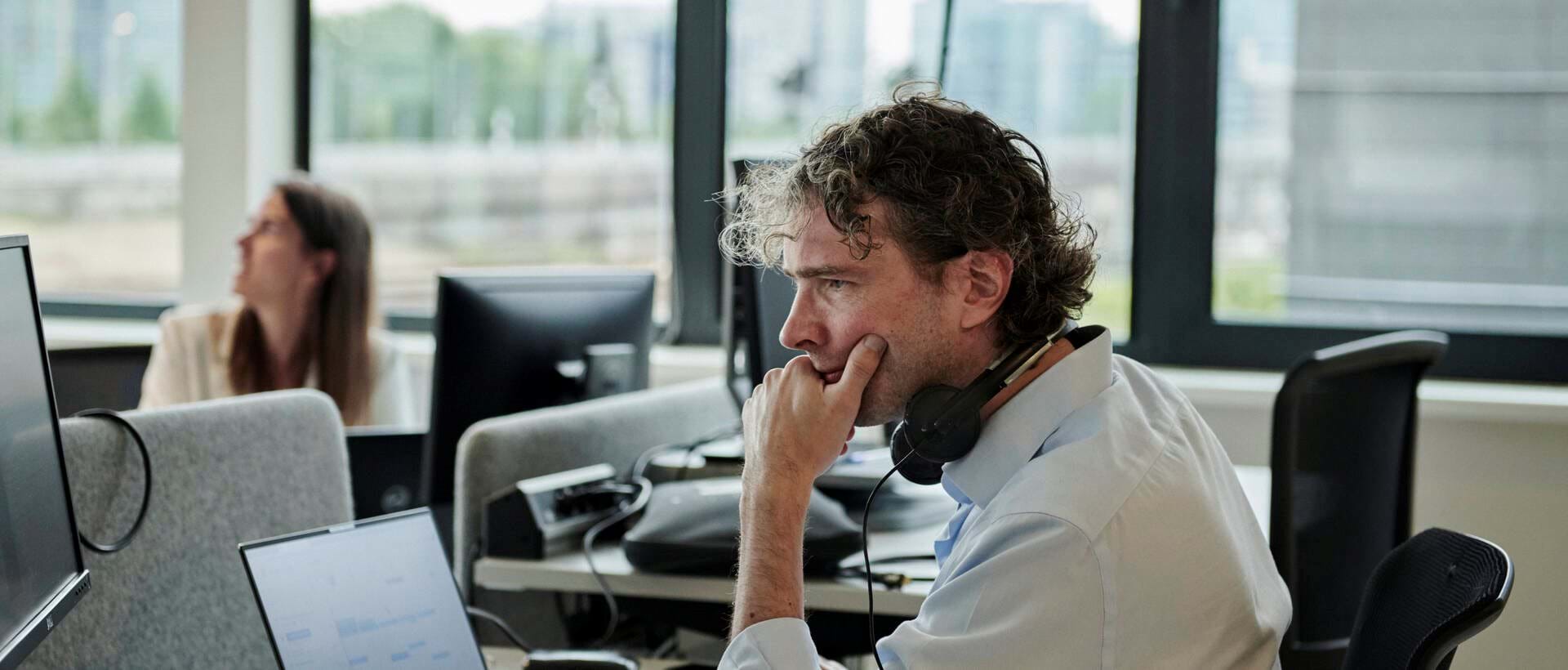 A man sits at a computer in an office by a big window, looking pensive.