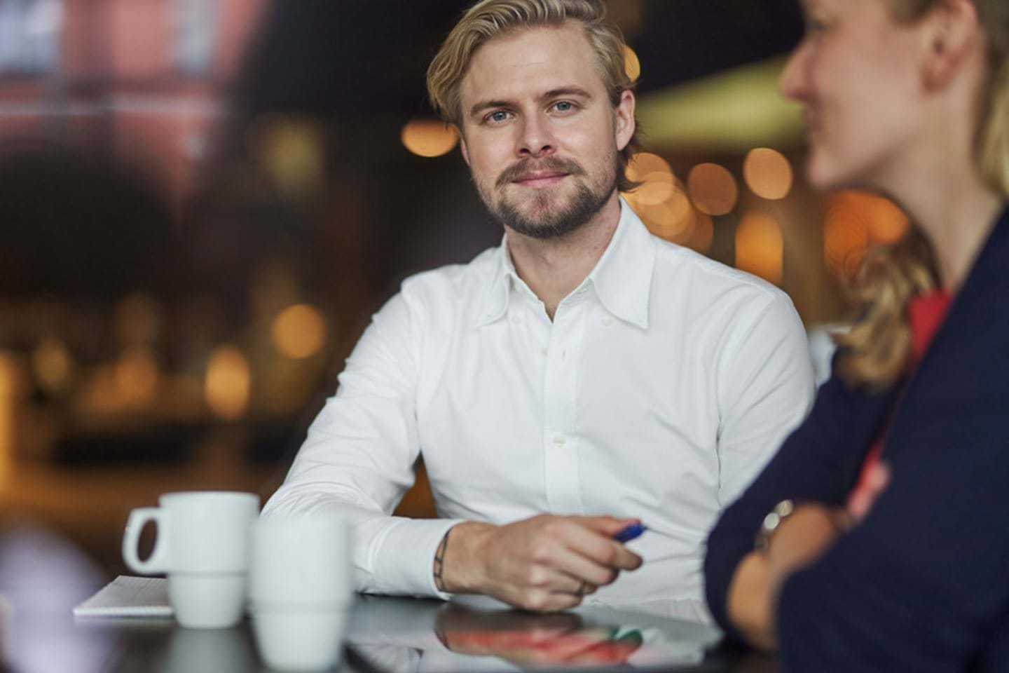 A man in a white shirt looks at the camera while sitting at a cafe table.