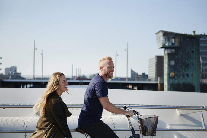 A woman sits on the back of a man's bike as they ride together over a bridge on a sunny day.
