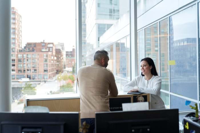 A man and woman have a conversation in the corner of the office next to a big window looking out over the city.