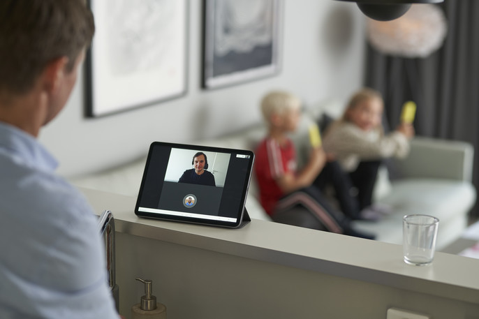 A man conducts a video call at home while his kids sit on the sofa in the background.
