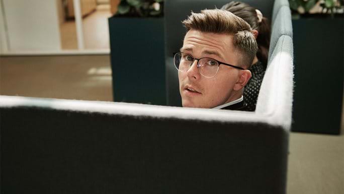 A young man with glasses looks up from a cosy bench corner at work.