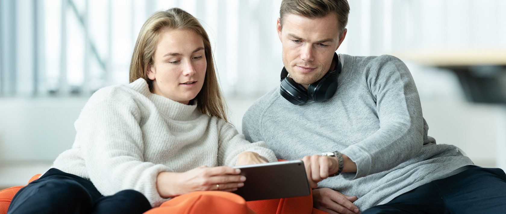 Woman in white sweater and man in grey sweater sitting in lounge chairs, leaning towards each other and looking at a tablet screen