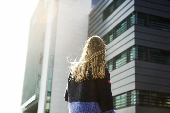 A blonde woman with long hair and her back to the camera walks towards a gray building illuminated by sunlight.