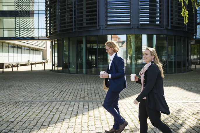 A professionally dressed woman and man carry coffee cups on a sunny day walking to their next meeting.