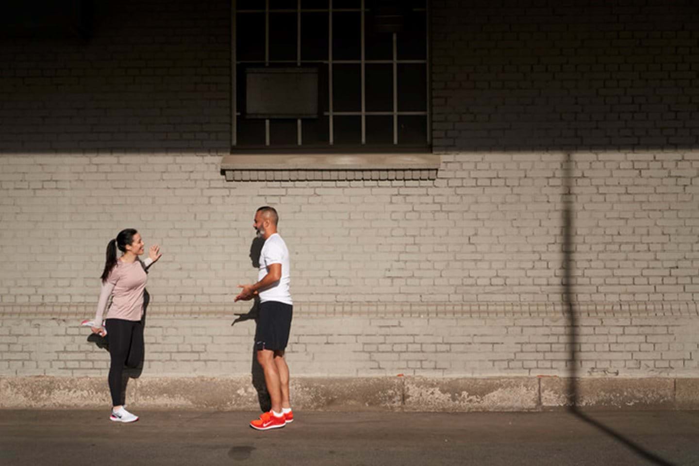 Two colleagues stretch on the sidewalk by a wall before a mid-day jog.