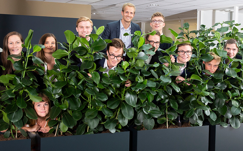 Management trainees joke around at work, posing for a photo behind some work plants.