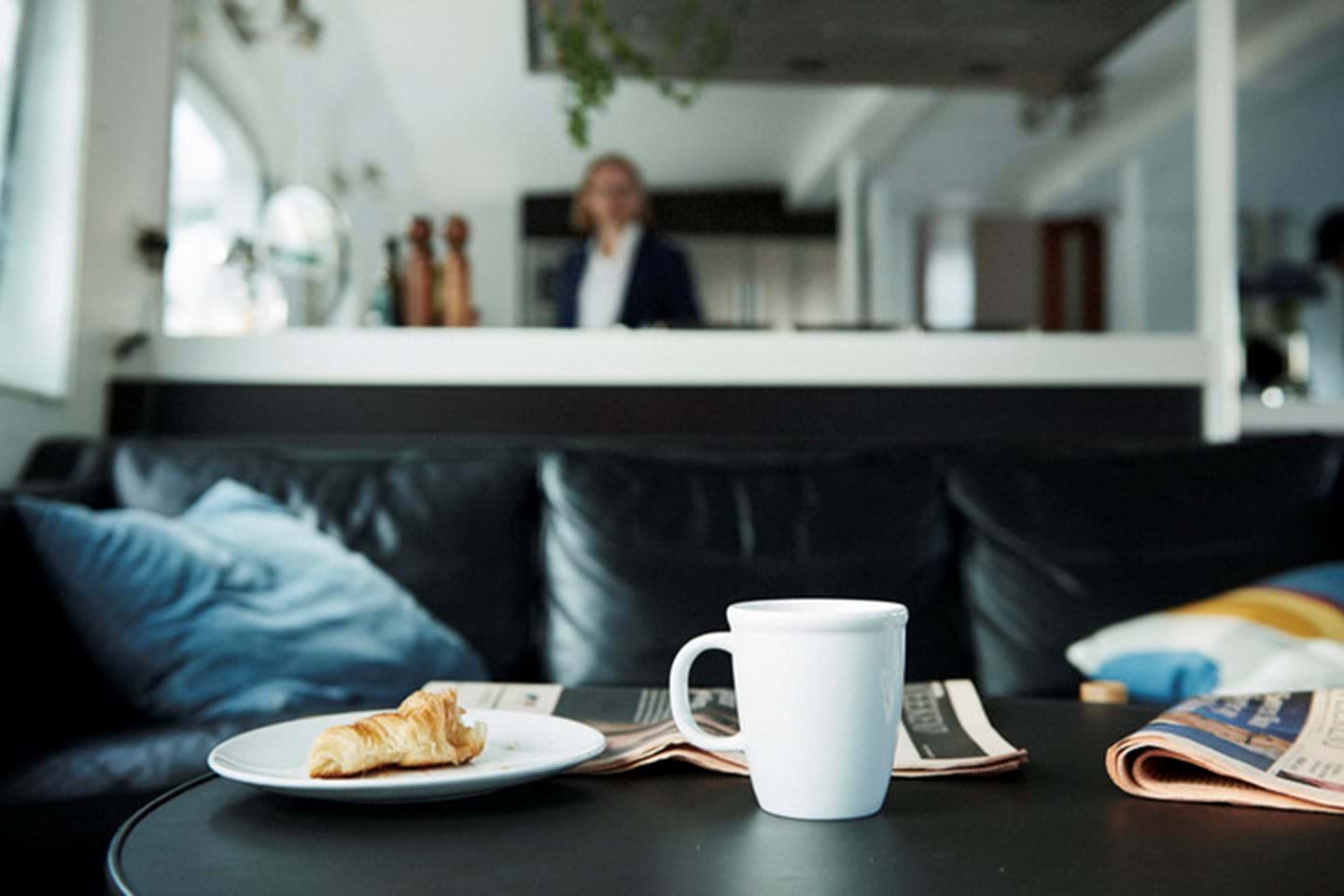 A man in a white shirt and jacket stands in his kitchen with his coffee and breakfast in focus.
