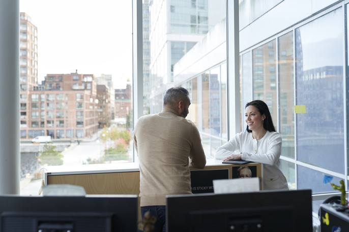 A man and woman have a conversation in the office next to a large window overlooking a city street.
