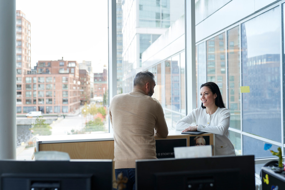 A woman chats with her male colleague at the office, overlooking a view onto the street.