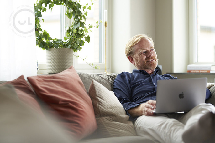 A man in a blue shirt sits on his sofa with his computer and looks into the distance.